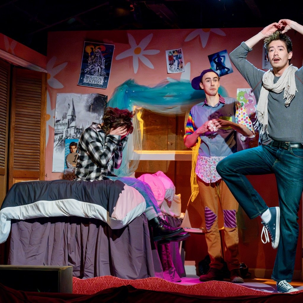 A young woman sits on a bed with her face in her hands while a young man poses on one leg with his hands over his head. Another man standing behind them looks on.