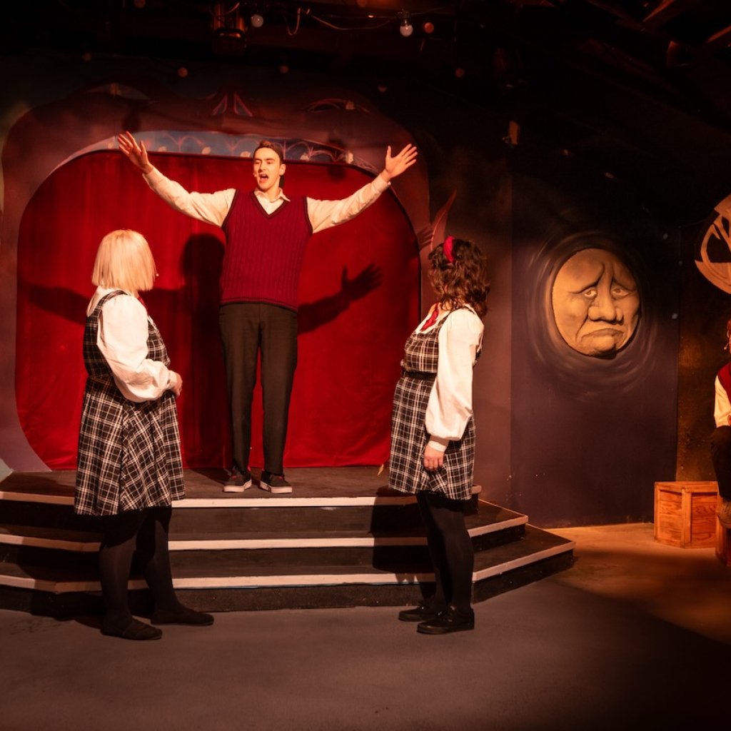 Archive photo of Ride the Cyclone. A man stands on an elevated stage with his arms raised in front of three other cast members.