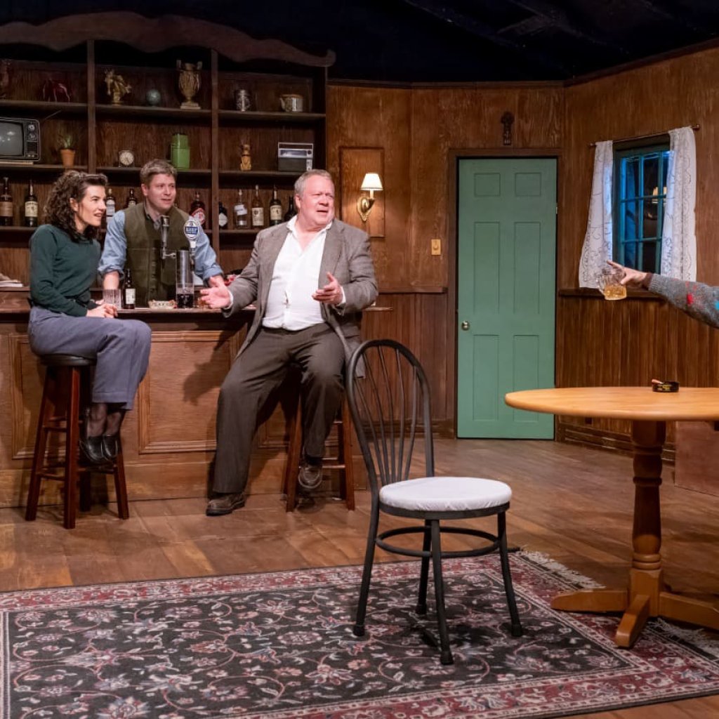 A seated pub patron raises his hand towards the bartender while other patrons look on