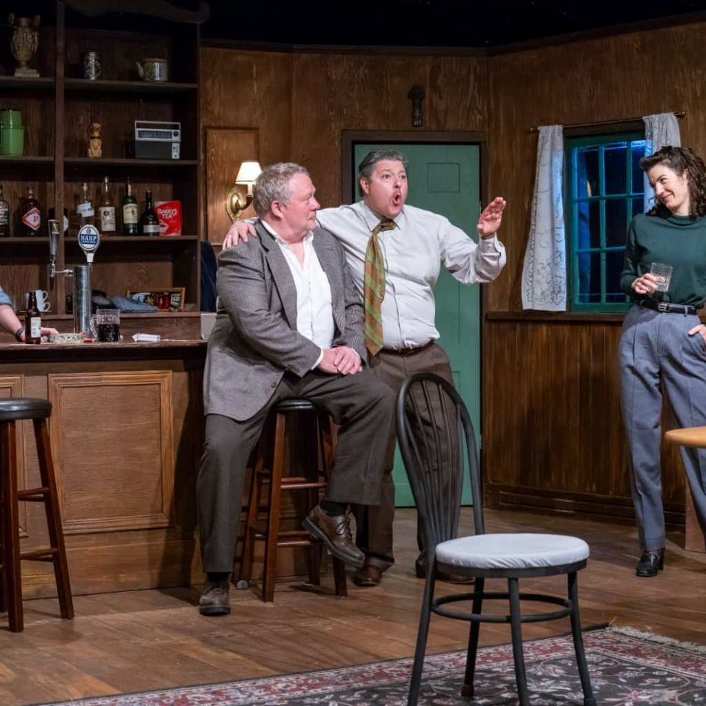 Four people in a pub. The man in the center has his arm on another man's shoulder as he gestures forward