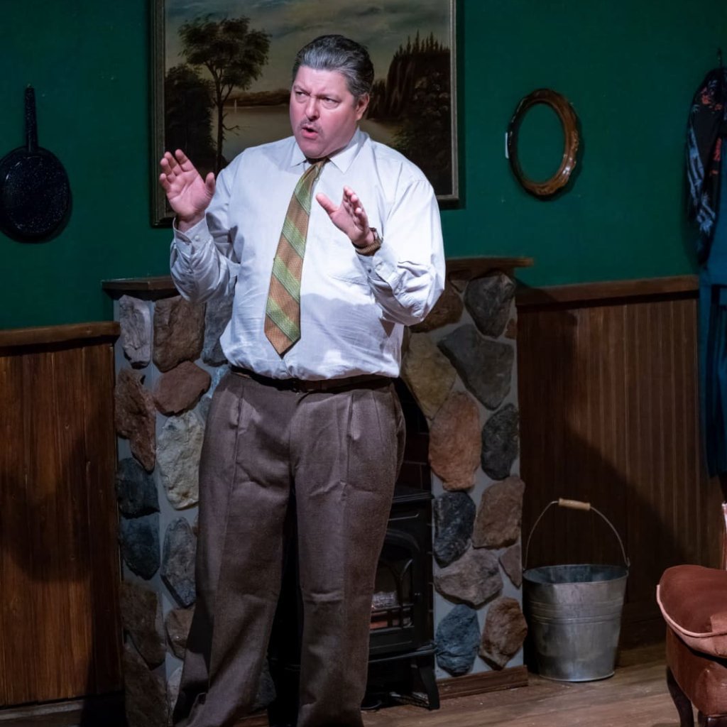 A man standing in front of a fireplace, speaking with his hands raised in front of him