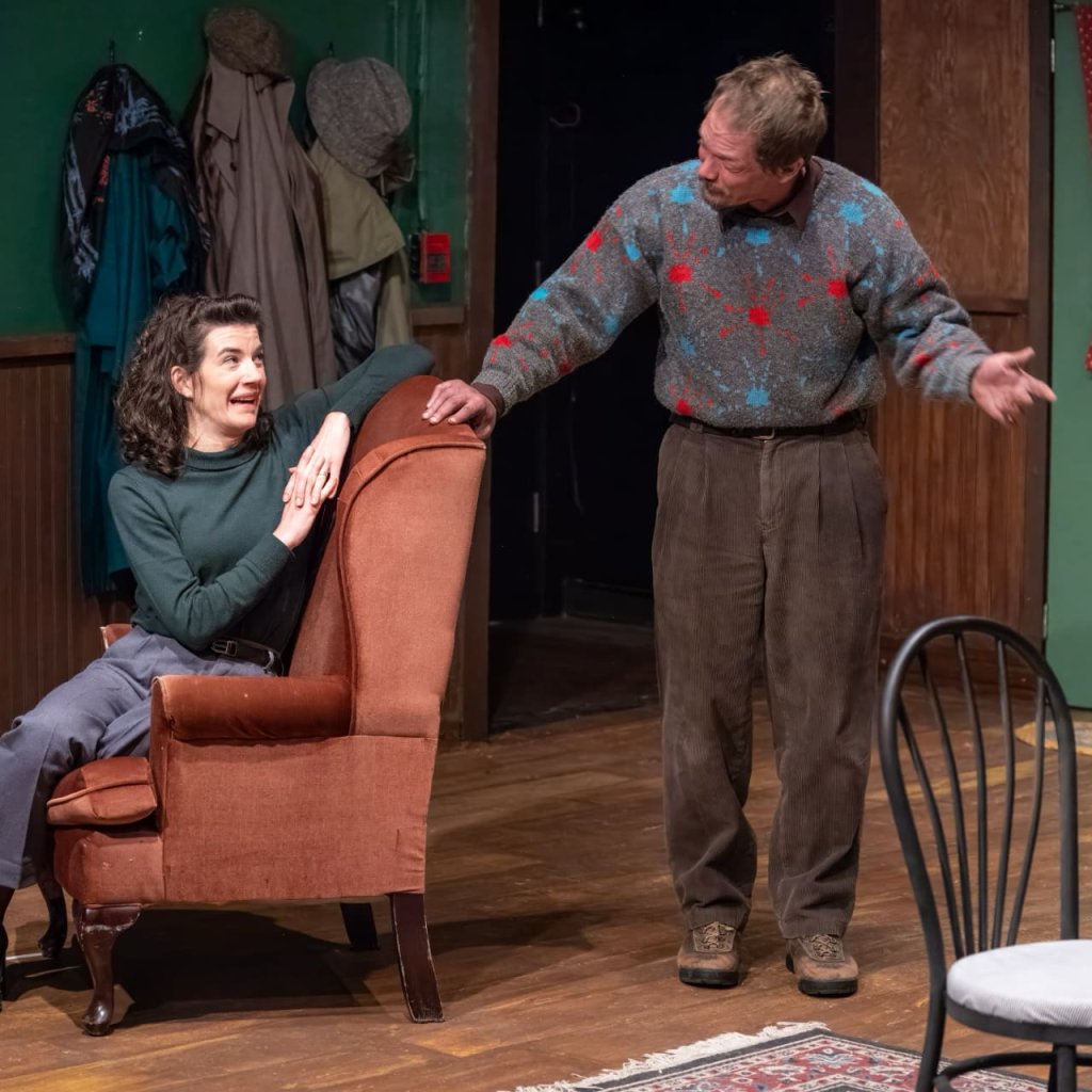 A standing man wearing a sweater speaks to a woman sitting in a red armchair