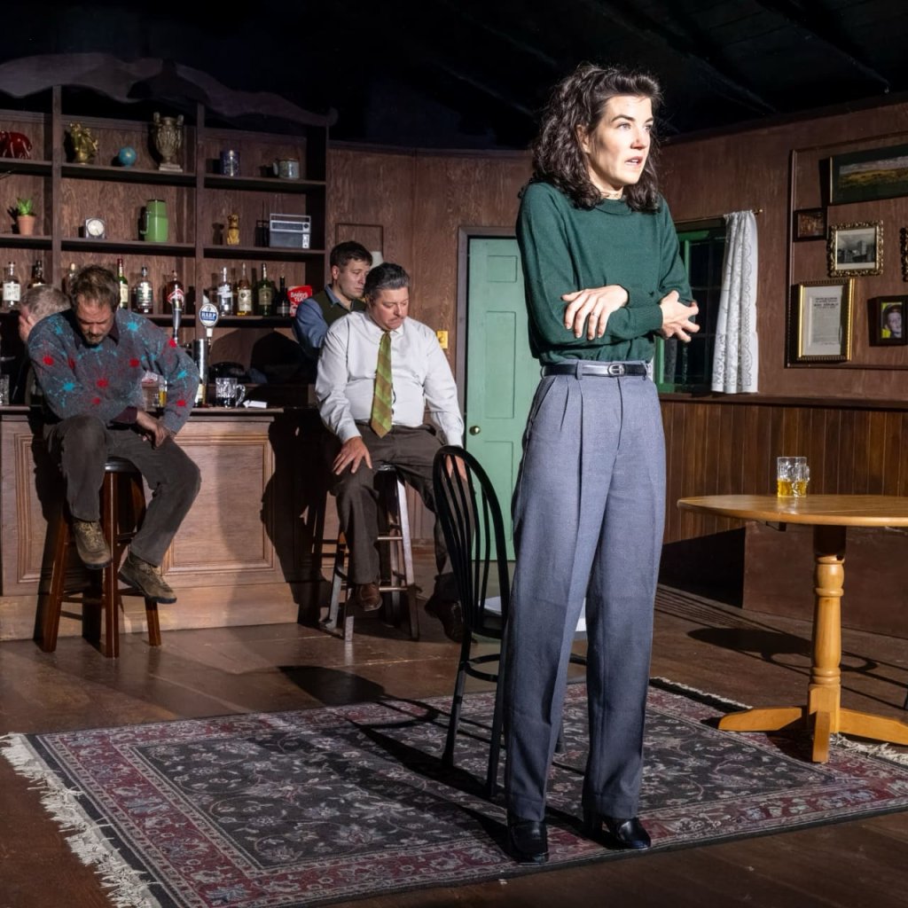 A woman with arms crossed stands in the middle of an Irish pub. Four men at the bar behind her have their heads lowered.