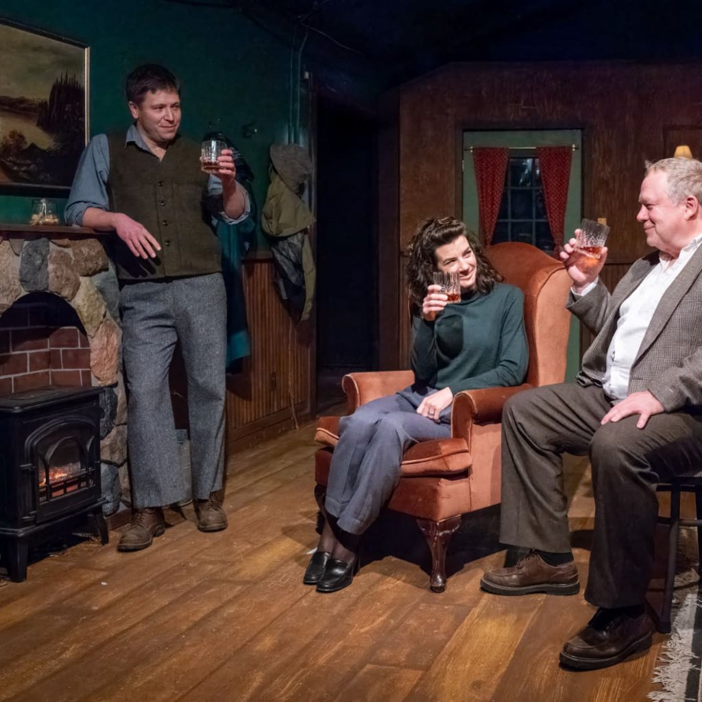 A man leans against a fireplace mantle while a woman and man sit by the fire. They raise their glasses to each other.
