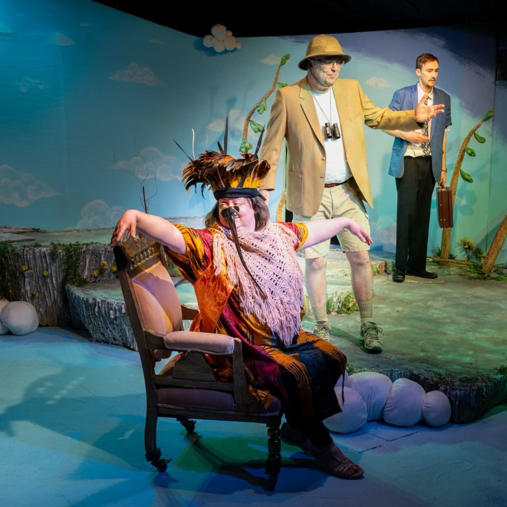 A woman in a feathered hat and a long black beak sits in a chair with arms spread. Two men stand in the background.