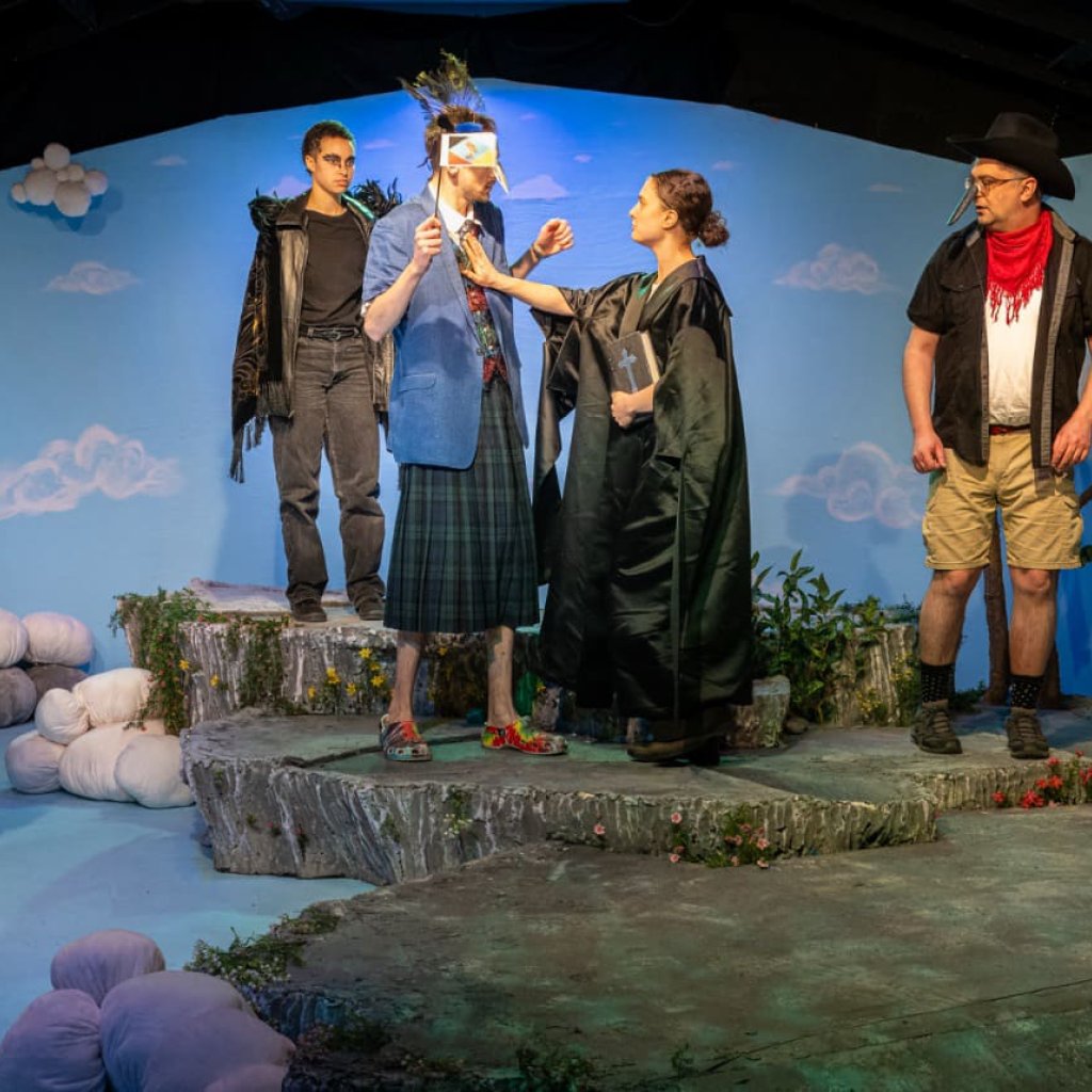 A woman dressed as a priest places her hand on a man's chest. He is dressed as a bird and holding a small flag.