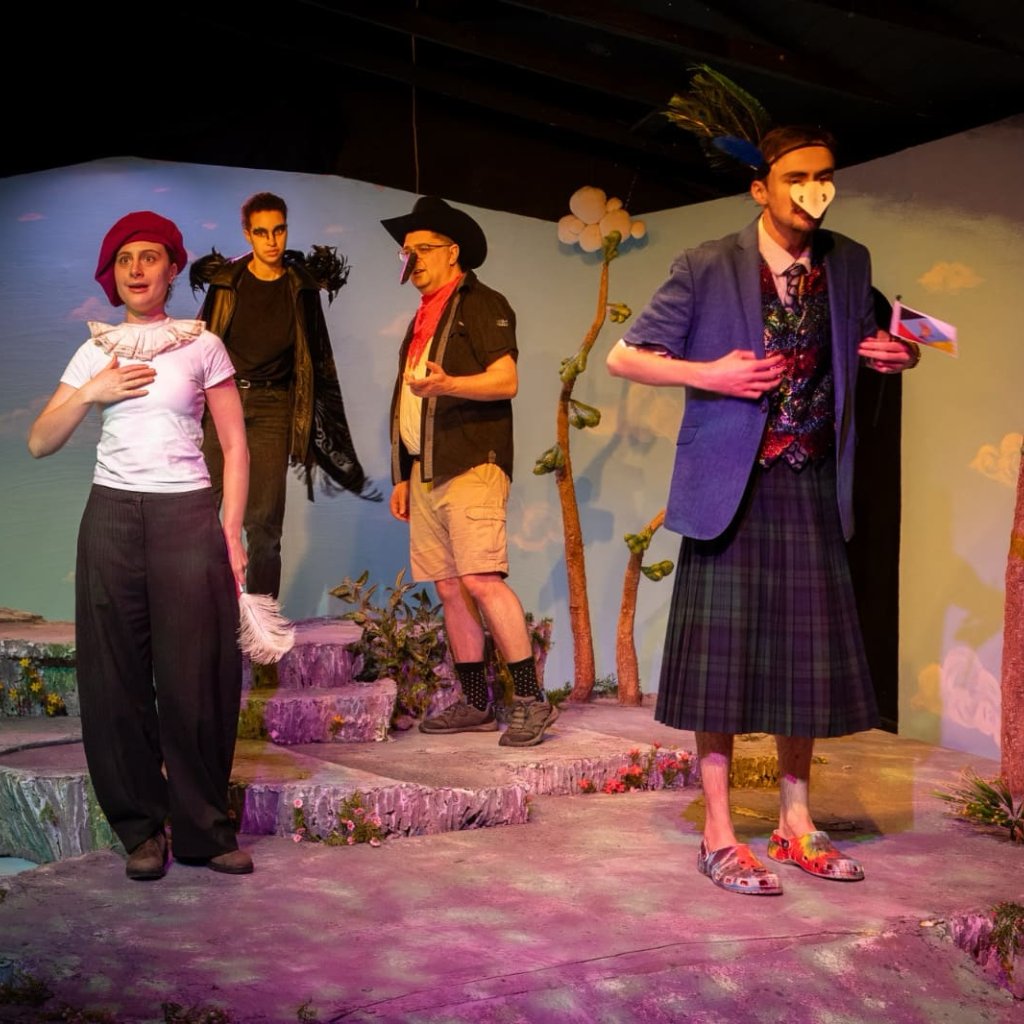 A woman speaks while holding a hand to her chest as three other people dressed as birds look on.