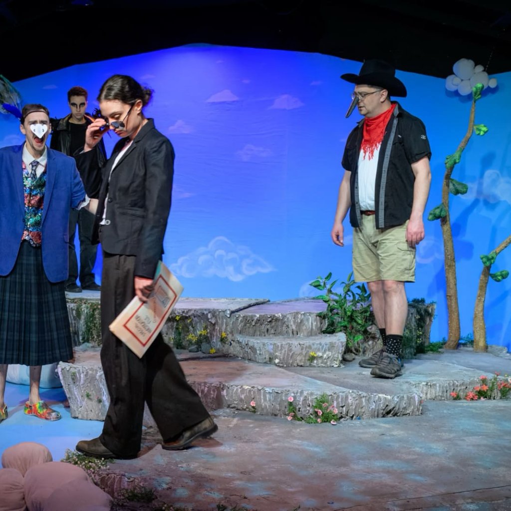 A woman wearing a suit looks over the top of sunglasses while a man dressed as a bird speaks animatedly to her